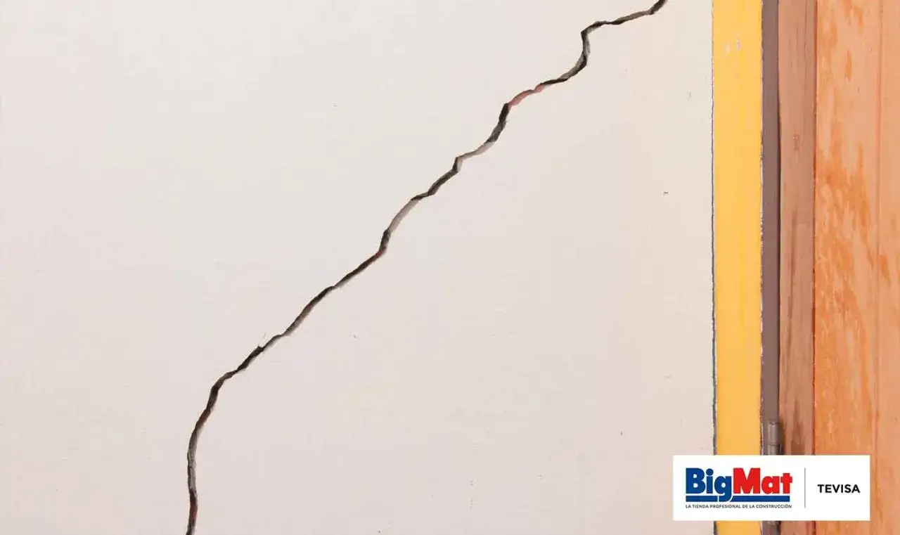 Grieta profunda en pared blanca, indicando la necesidad de reparar grietas por asentamiento. Se ve un marco amarillo y madera.
