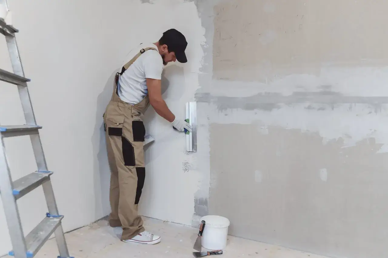 Obrero con mono de trabajo realiza el enfoscado de yeso en una pared, con una escalera a un lado y herramientas en el suelo.