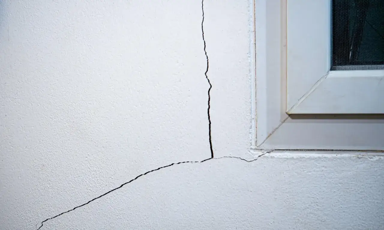 Tipos de grietas en paredes: una grieta fina y otra más gruesa se extienden desde la esquina de una ventana blanca hacia el interior de la pared.