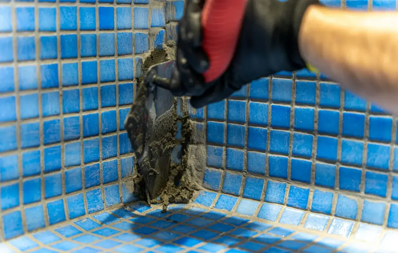 Mano enguantada aplica mortero para impermeabilizar piscina con mosaicos azules.