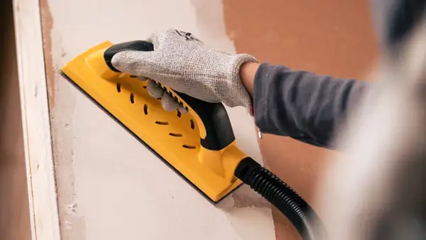 Mano enguantada usando una lijadora amarilla para lijar paredes, preparando la superficie para pintar.