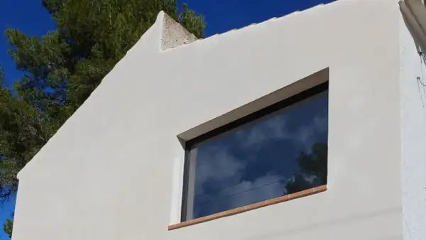 Fachada blanca con mortero de cal, reflejando el cielo azul y nubes. Ventana moderna y árbol verde.