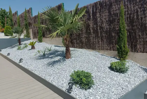 Un jardín moderno con palmeras y cipreses, decorado con piedras blancas y luces integradas.