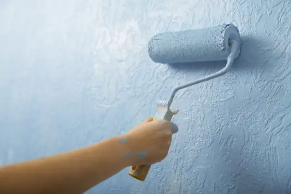 Mano pintando una pared con rodillo azul. El plástico del rodillo se ve salpicado de pintura.