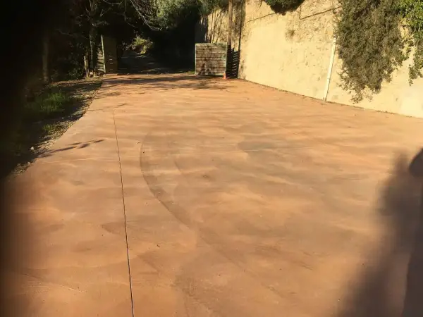 Un camino de **suelo hormigon pintado** de color terracota, con marcas de neumáticos, conduce a una puerta abierta.