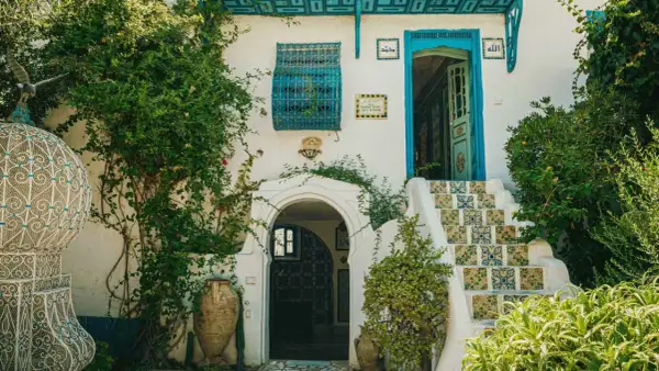 Fachadas antiguas con escaleras de azulejos coloridos, puerta azul y vegetación exuberante. Un portal arqueado y vasijas de barro completan la escena.