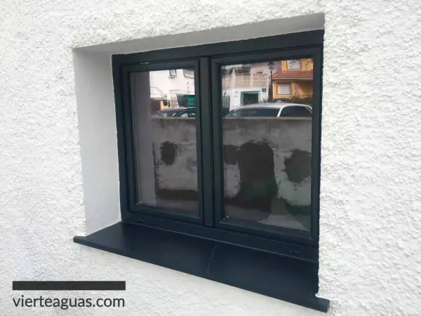 Ventana con marco oscuro y alféizar negro, lista para rematar ventanas por fuera. Refleja una calle con casas y coches.